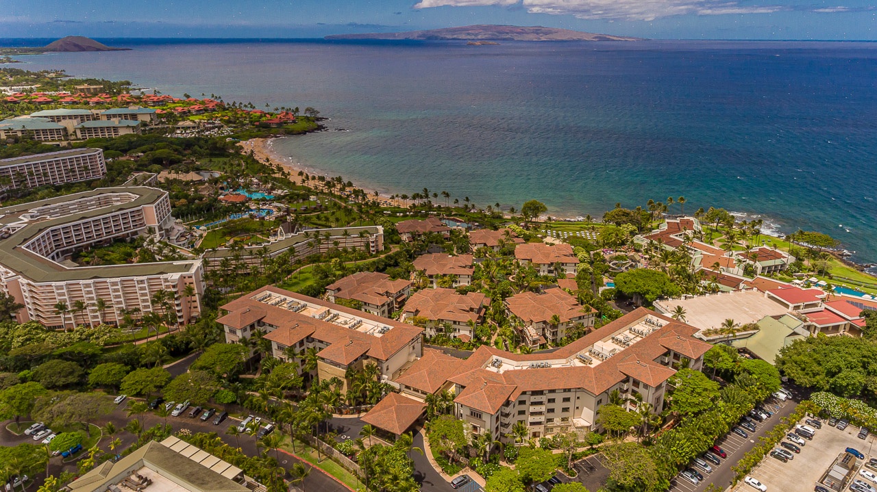 An aerial view captures the beachfront property nestled along a pristine coastline, surrounded by lush tropical landscaping and neighboring resort accommodations.