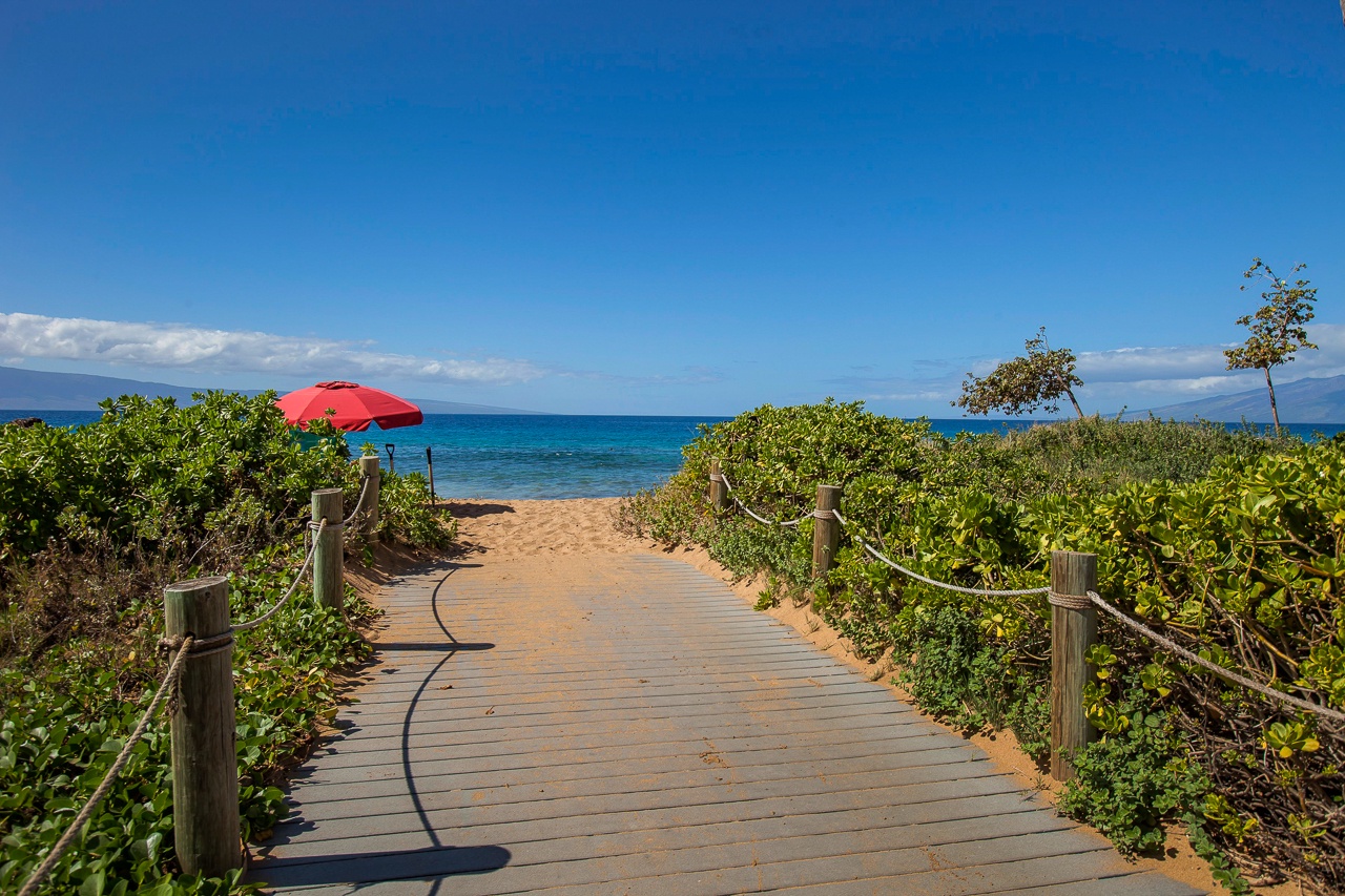 A wooden boardwalk leads through coastal greenery to golden sand and calm turquoise waters,.