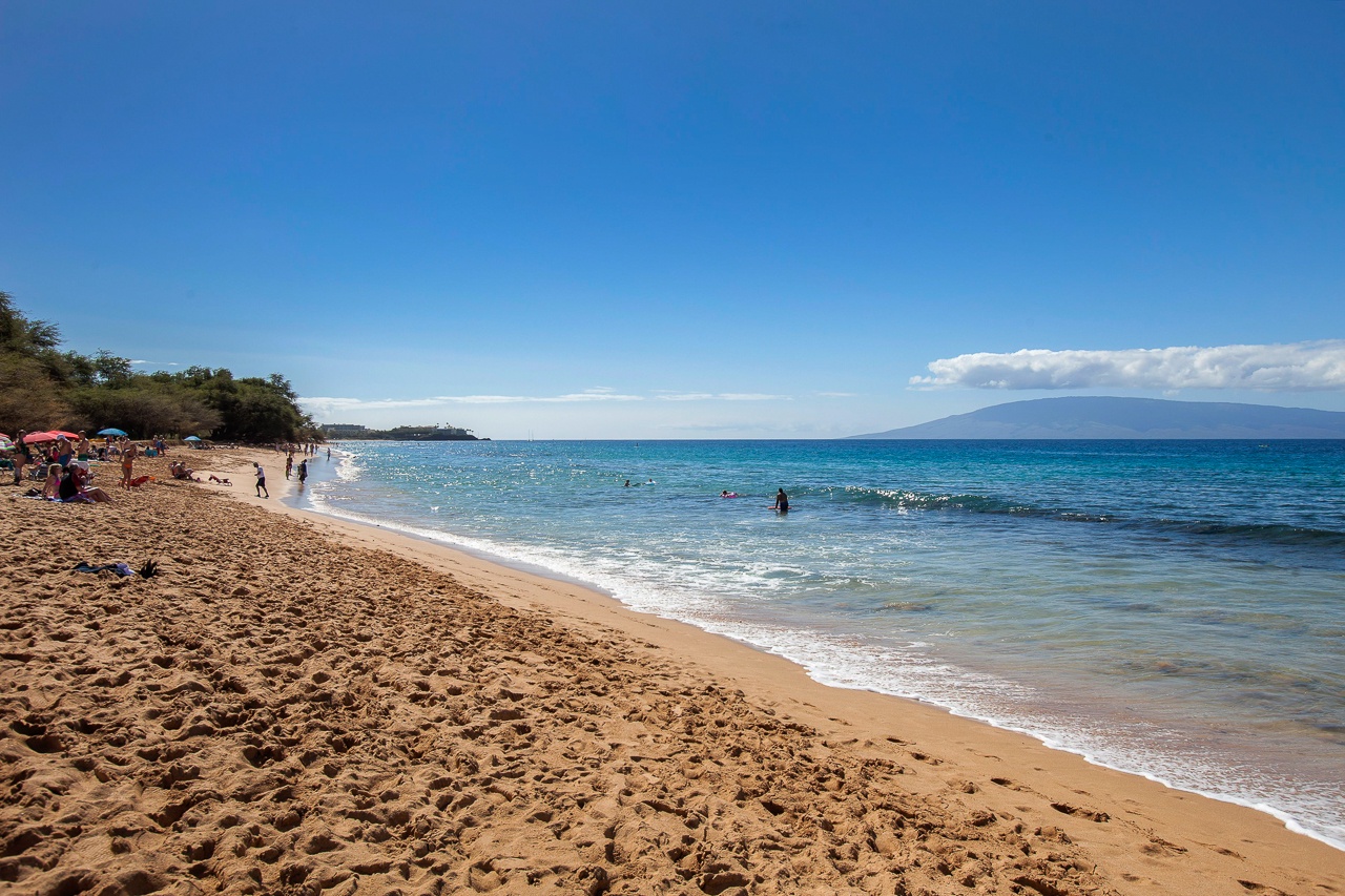 A beautiful sandy beach stretches along sparkling turquoise waters, with gentle waves rolling in under a clear blue sky and distant mountain views on the horizon.