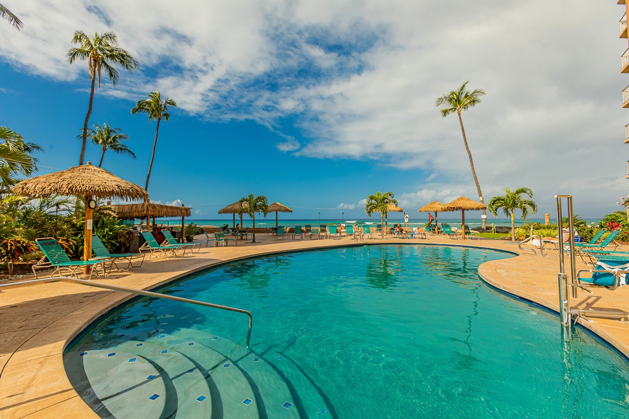 Resort-style pool with thatched palapas framed by swaying palms, set against turquoise ocean waters and open skies. An inviting tropical oasis awaits your arrival.