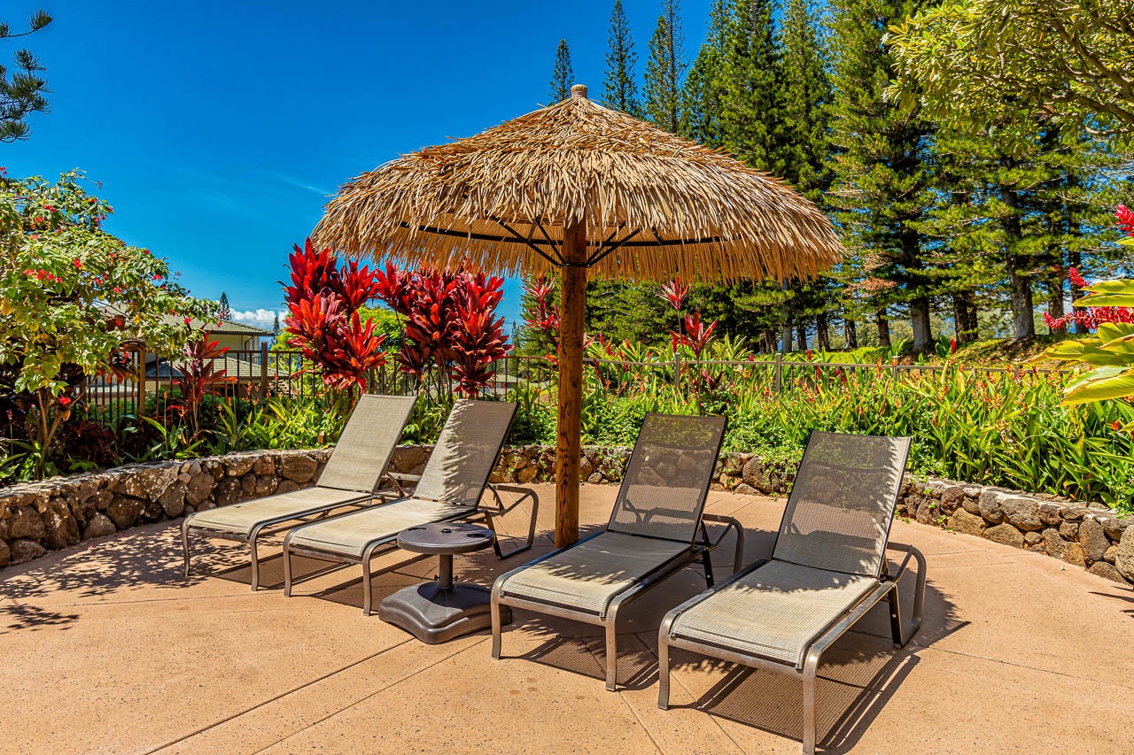 Four cushioned loungers await beneath a thatched palapa, surrounded by vibrant tropical blooms and swaying pines under endless blue skies—your perfect escape.