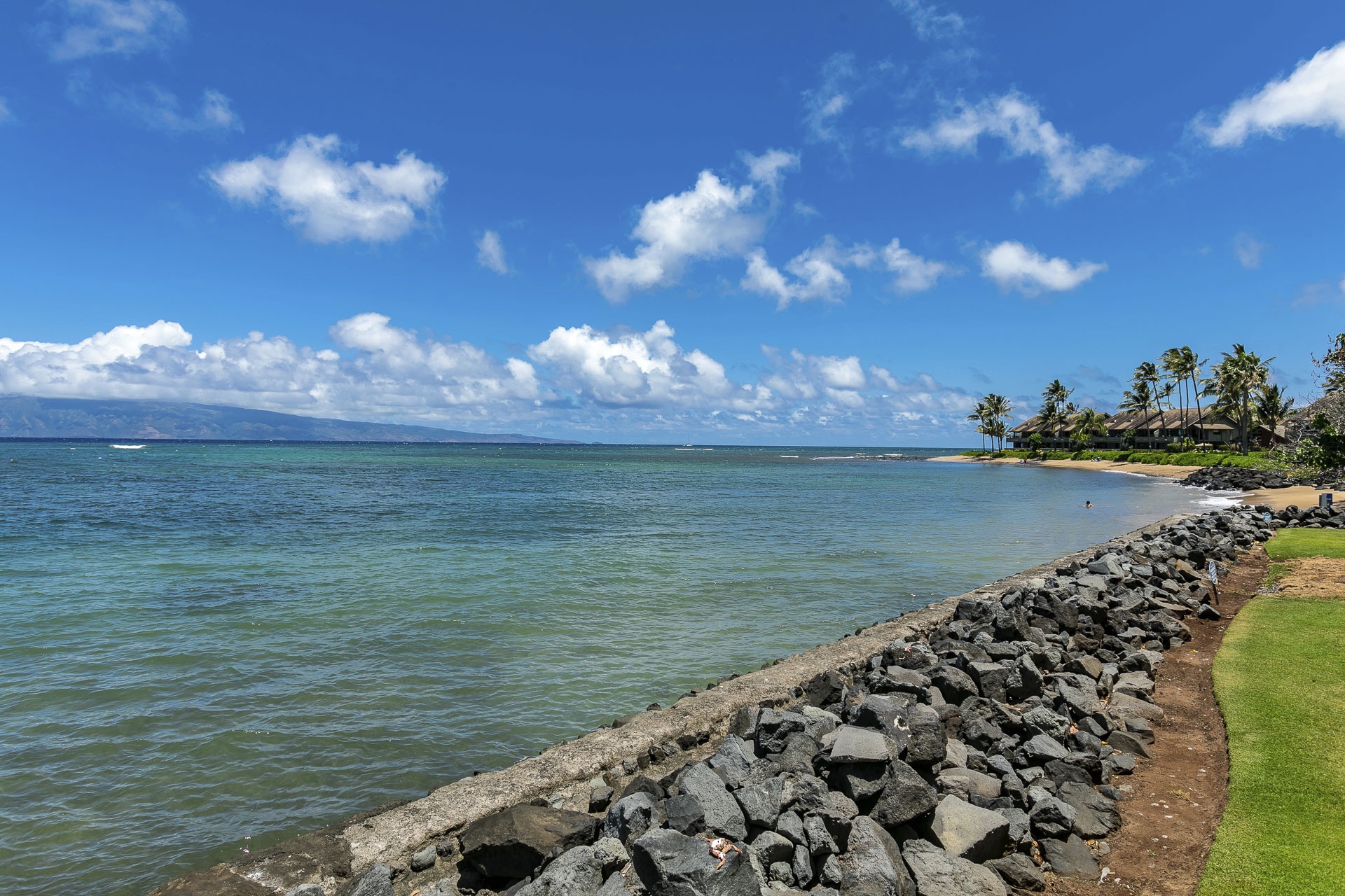 Tranquil shoreline with volcanic rock lining, sandy beaches, and palm-fringed coast stretching along the calm waters.