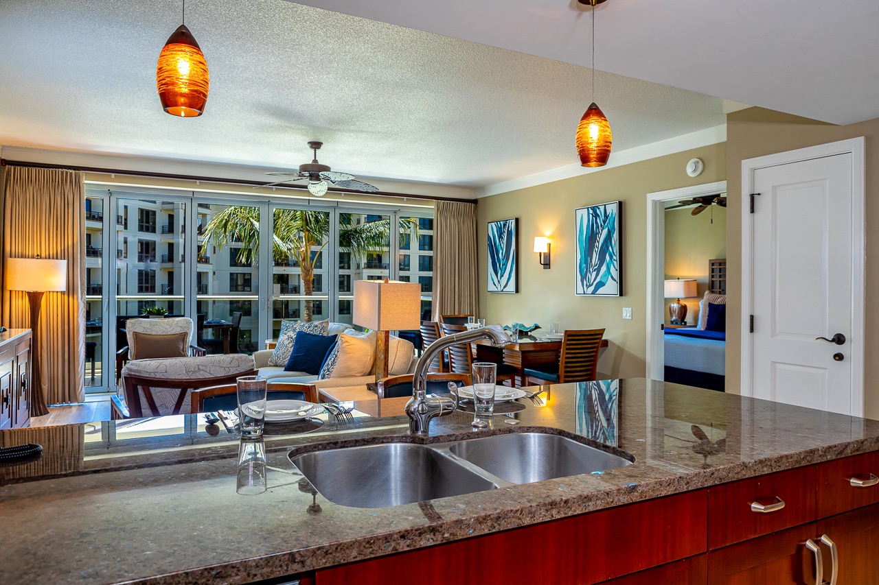 Bright, open living space with fully-equipped kitchen flowing into dining and lounge areas. Floor-to-ceiling glass doors frame tropical views and flood the room with natural light.