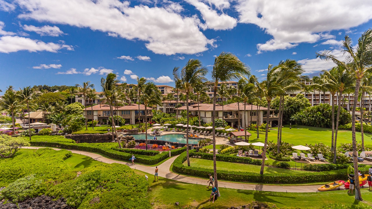 Aerial view of a tropical resort property with manicured gardens, swaying palms, and a central swimming pool area nestled among low-rise buildings.