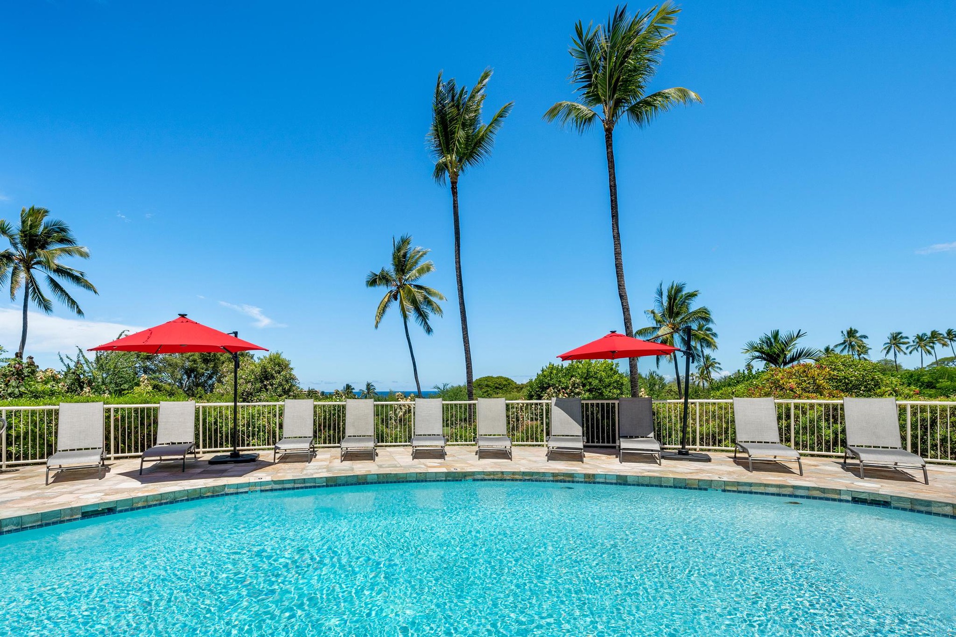 Crystal-clear pool with tropical palms swaying overhead, surrounded by comfortable loungers and vibrant red umbrellas for perfect shade.