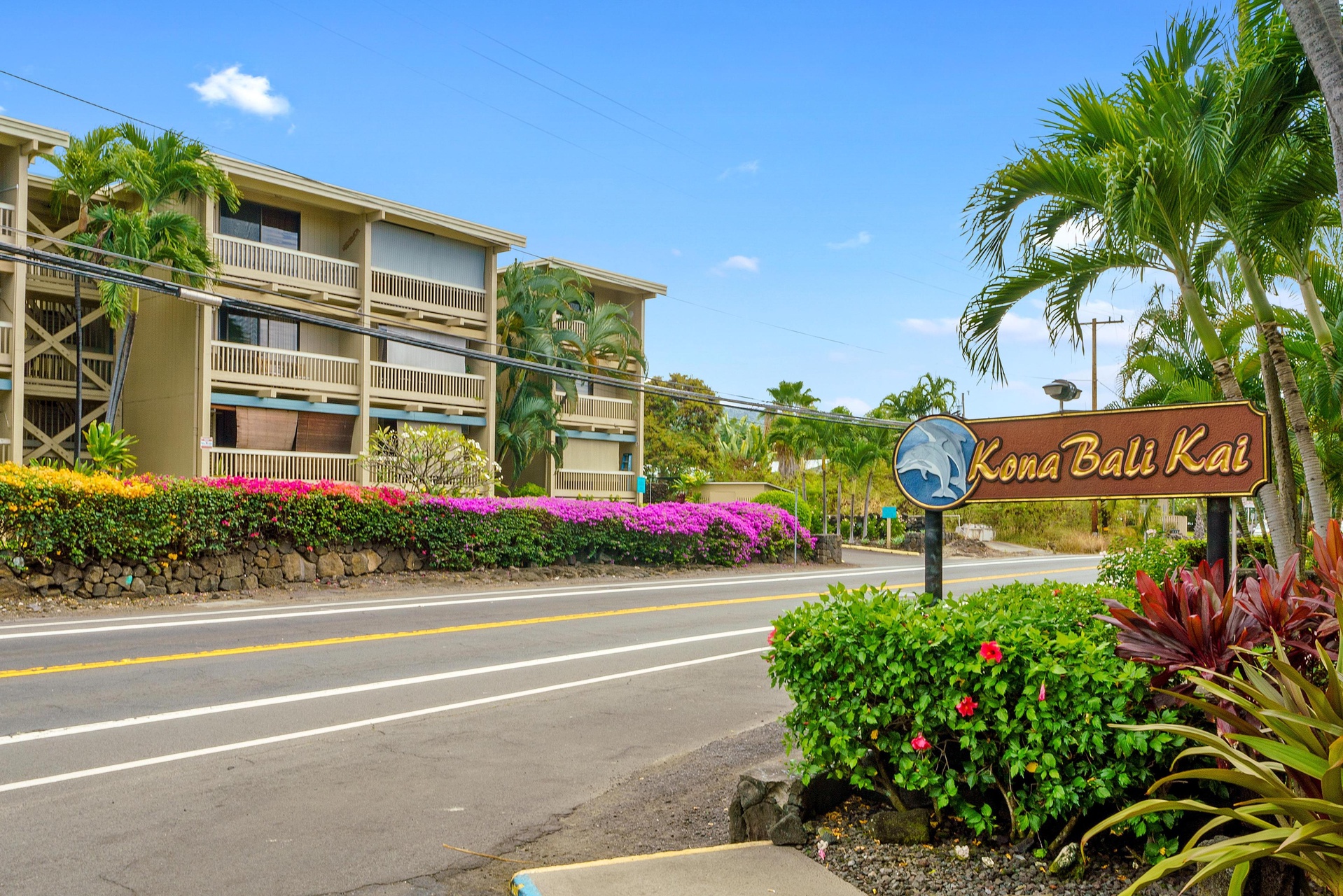 The property entrance is marked by vibrant tropical landscaping and the welcoming Kona Bali Kai sign along a well-maintained street under clear blue skies.