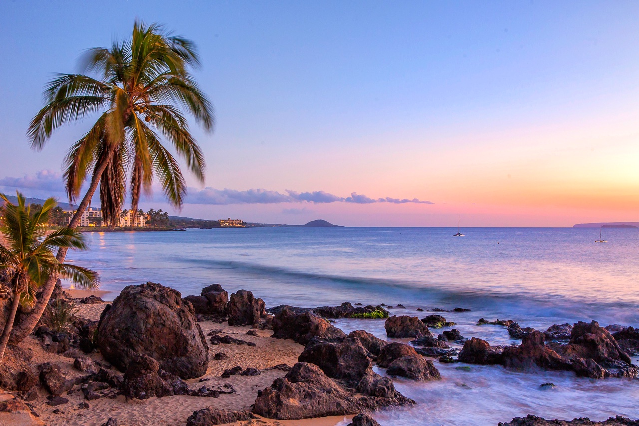 Tropical paradise with swaying palms and gentle waves lapping volcanic rocks creates the perfect sunset backdrop for your Hawaiian getaway.