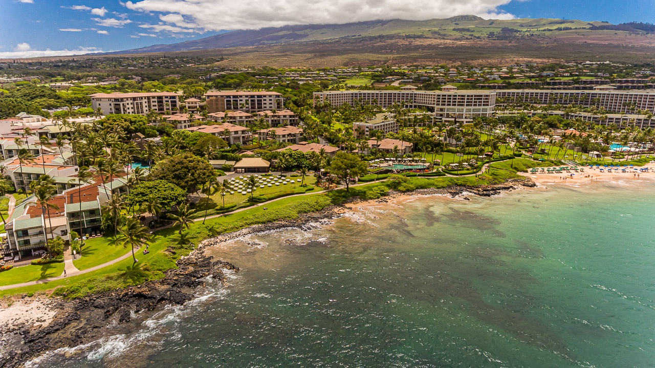Aerial view of a beachfront resort complex nestled along tropical shores, with majestic mountain slopes rising in the distance and lush green grounds stretching along the coastline.