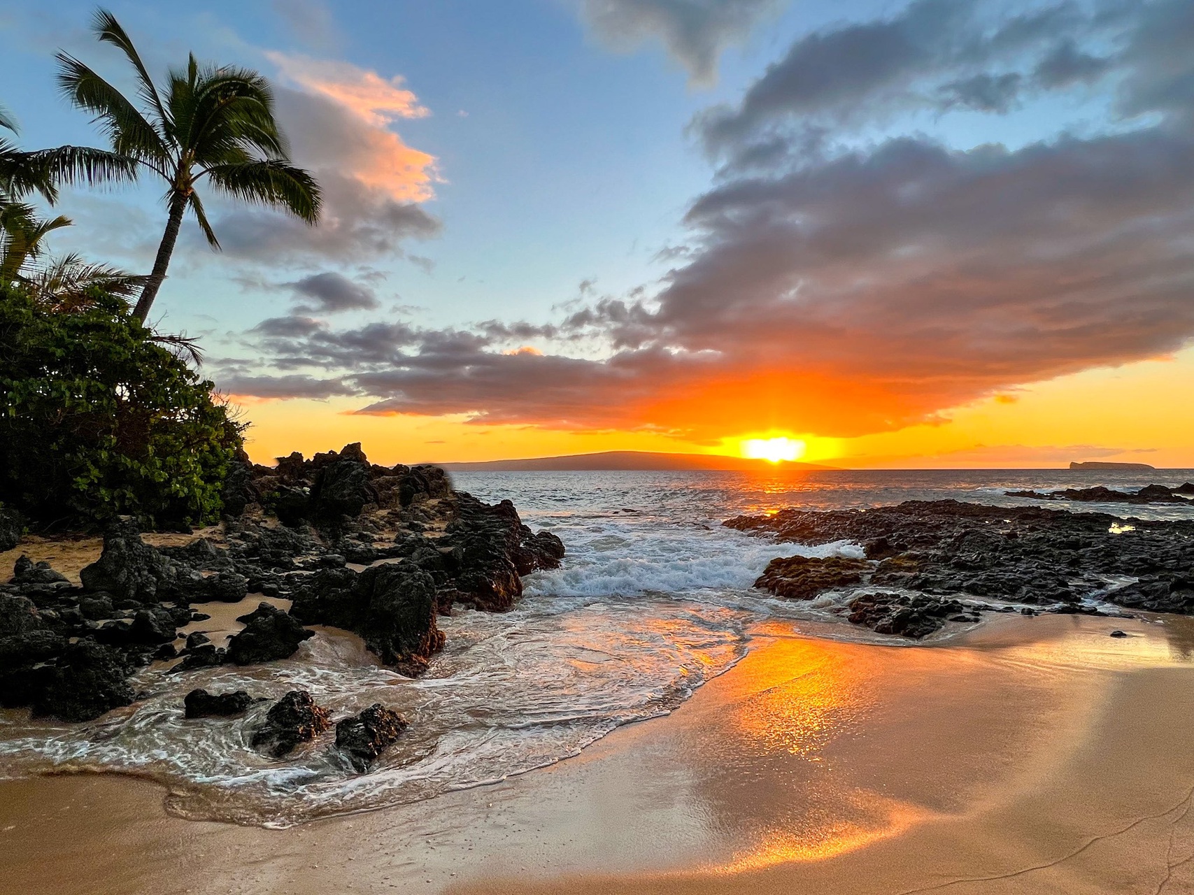A breathtaking tropical sunset paints the sky in vivid oranges and golds over a secluded beach, framed by swaying palms and dramatic volcanic rock formations along the shoreline.
