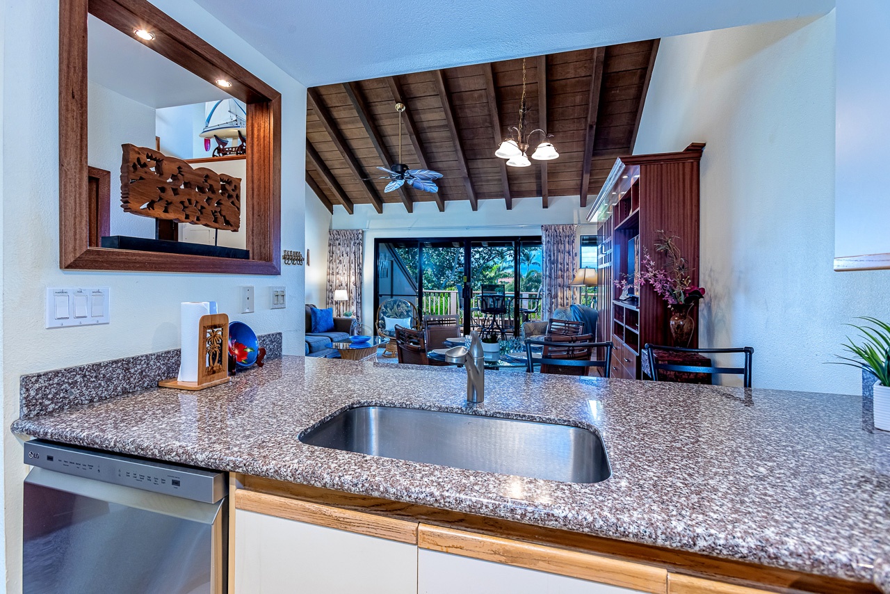 Step into your island escape where vaulted wood-beam ceilings and an open layout invite you to unwind. From this kitchen, the living area flows seamlessly to your private lanai, framed by lush greenery just beyond.