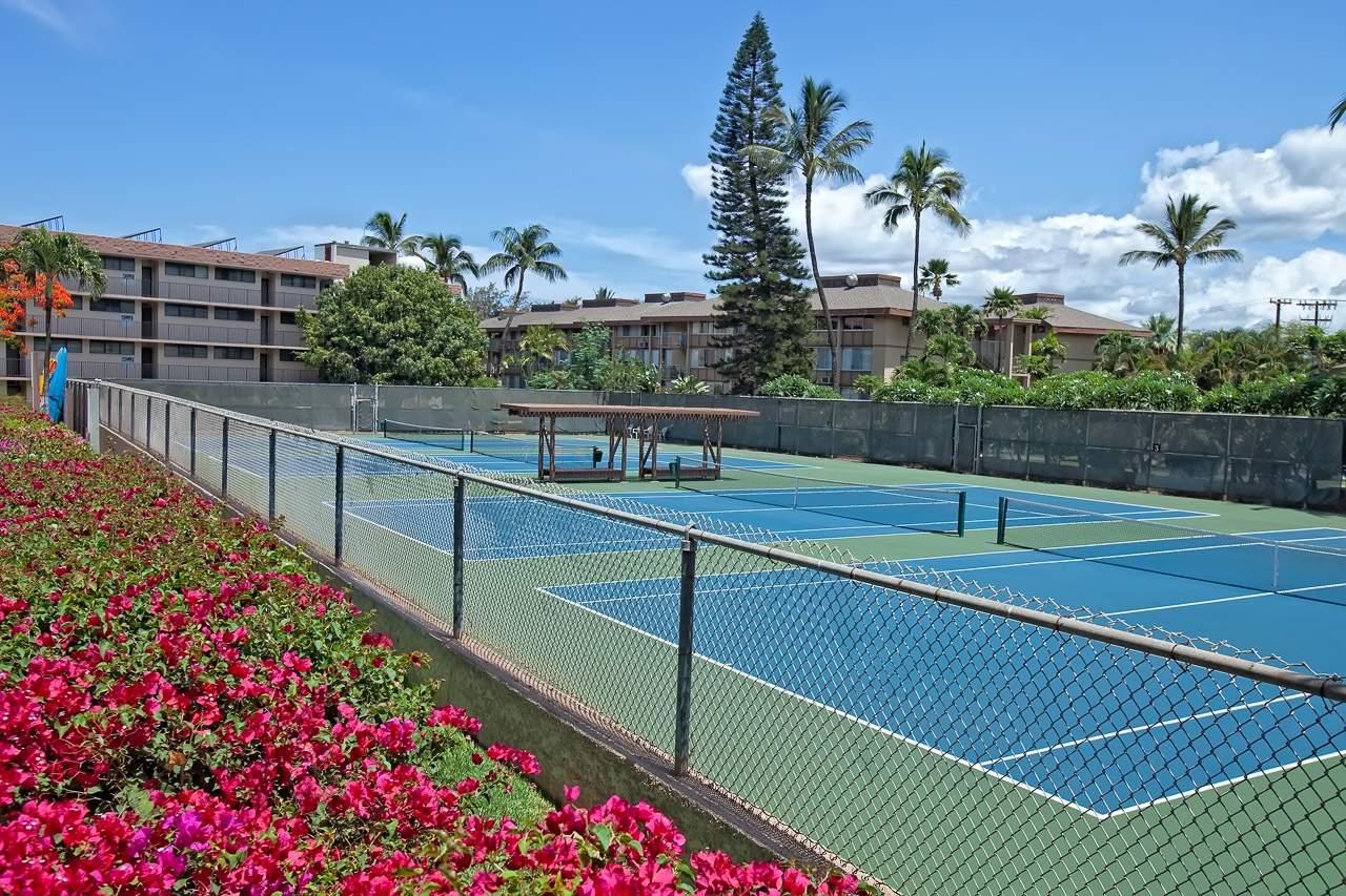 The property's tennis courts await your match, framed by vibrant tropical blooms and swaying palms under clear Hawaiian skies.