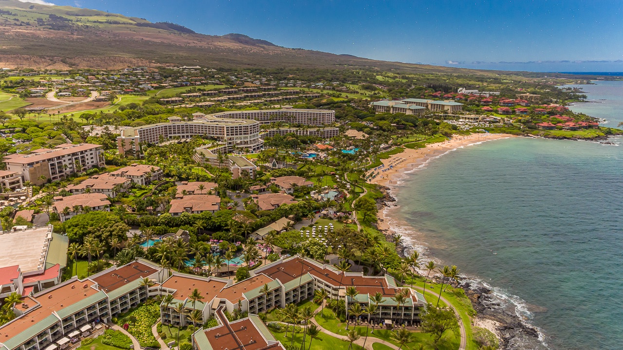 Aerial view of a stunning beachfront resort nestled along the coast, with sprawling grounds, multiple pools, and direct beach access—all set against majestic mountain slopes and clear blue waters.