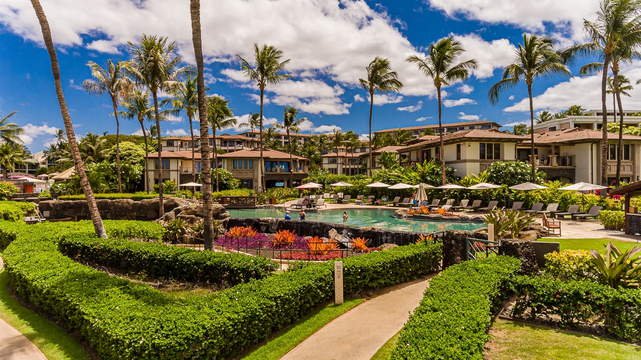 The resort features lush tropical gardens with manicured lawns and colorful plantings, a pristine swimming pool surrounded by comfortable lounge chairs and white umbrellas, all framed by swaying palm trees and blue skies.