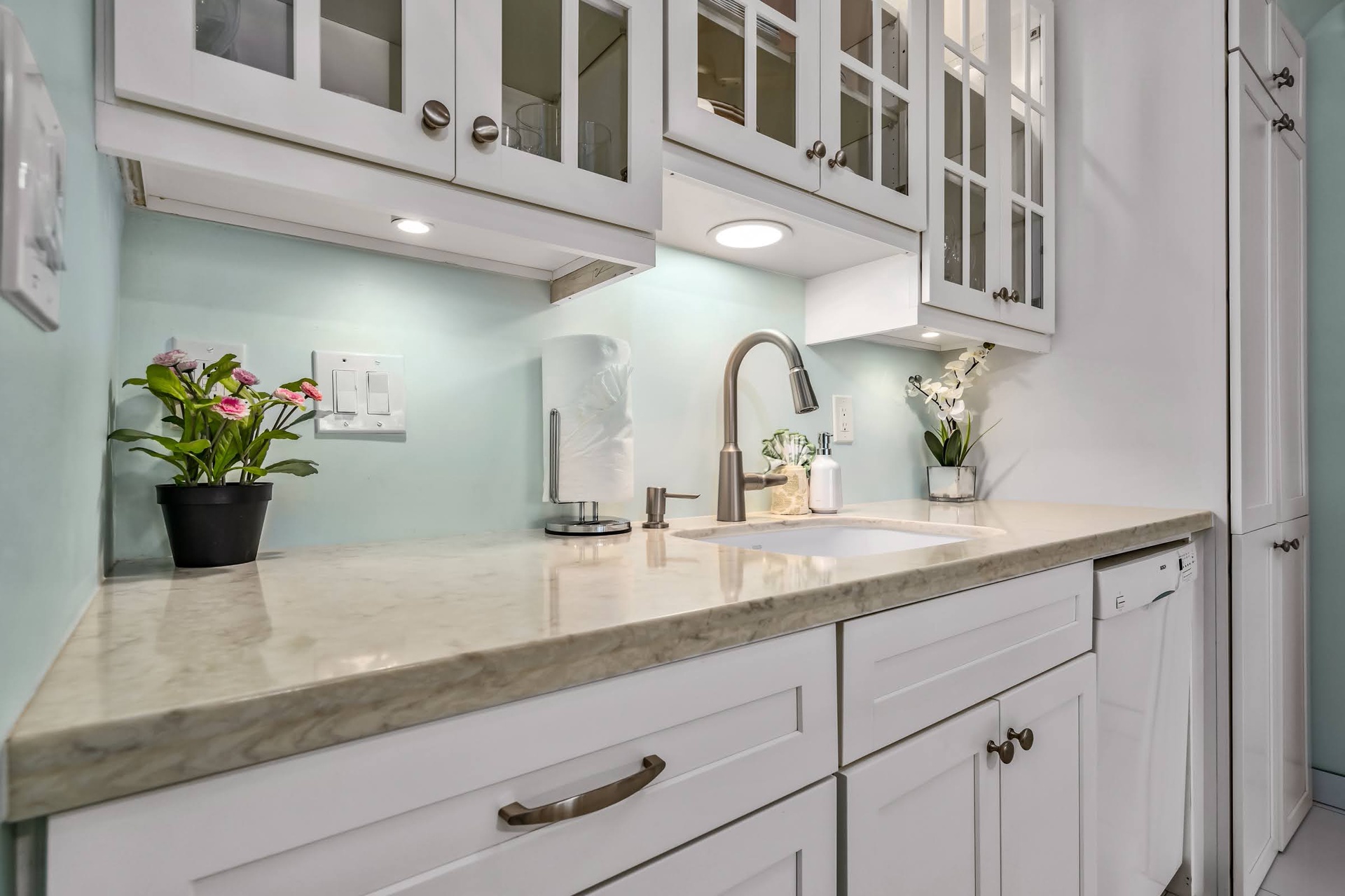 Prepare your favorite meals in this beautifully appointed kitchen featuring sleek marble countertops, soft under-cabinet lighting, and elegant glass-front cabinetry. Fresh details like potted flowers add a welcoming touch.