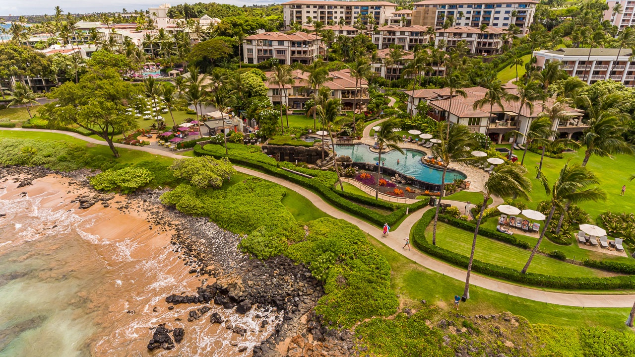 Beachfront resort property nestled along tropical shoreline, featuring landscaped grounds with pool and walking paths amid swaying palms and nearby accommodations.