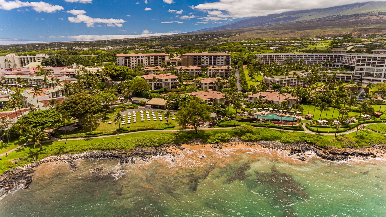 An aerial view captures the property's stunning beachfront location, nestled between turquoise waters and a lush resort complex surrounded by tropical landscaping and mountain vistas.