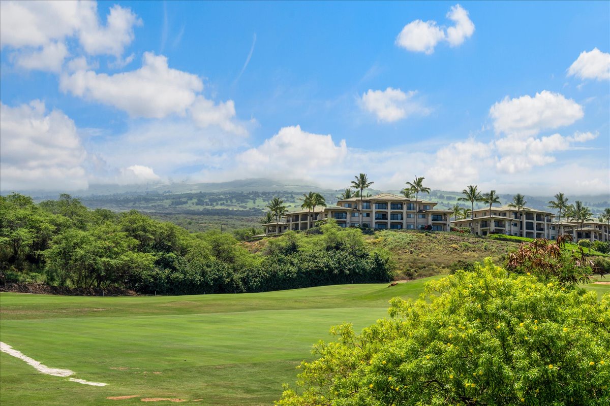 A resort property set amid tropical landscaping, surrounded by manicured greens, lush vegetation, and distant mountain views beneath a bright blue sky.