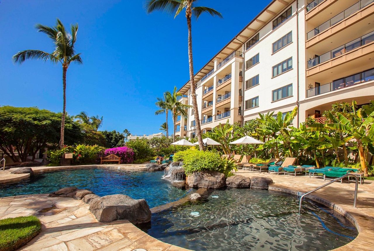 The resort building stands alongside a tropical pool surrounded by lush palms and vibrant greenery under brilliant blue skies.