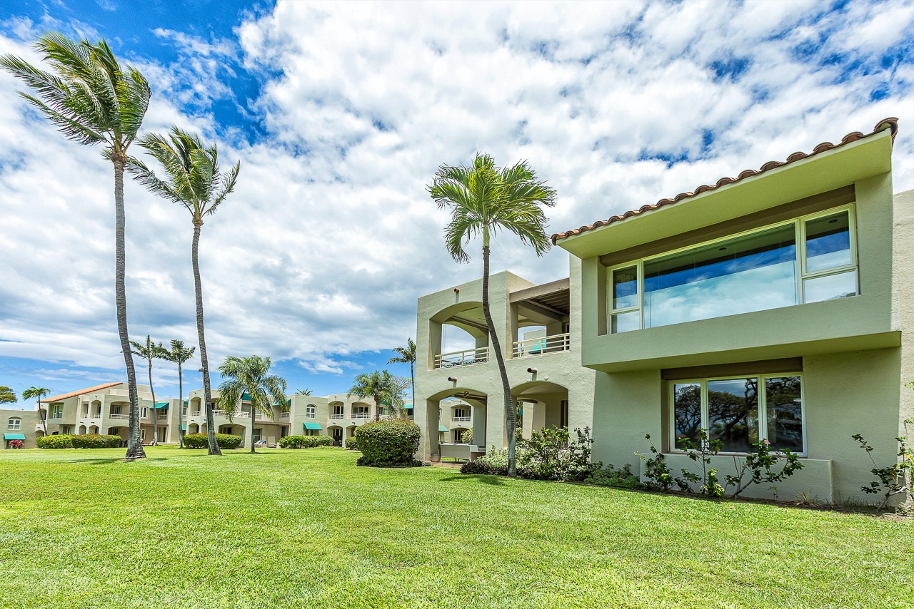 A tropical resort building set against lush green lawns and framed by swaying palm trees under a bright blue sky.