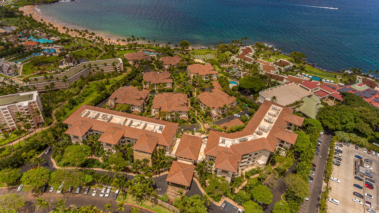 Aerial view of a beachfront resort complex with tile-roofed buildings, lush tropical landscaping, swimming pools, and direct access to sandy shores and calm ocean waters.