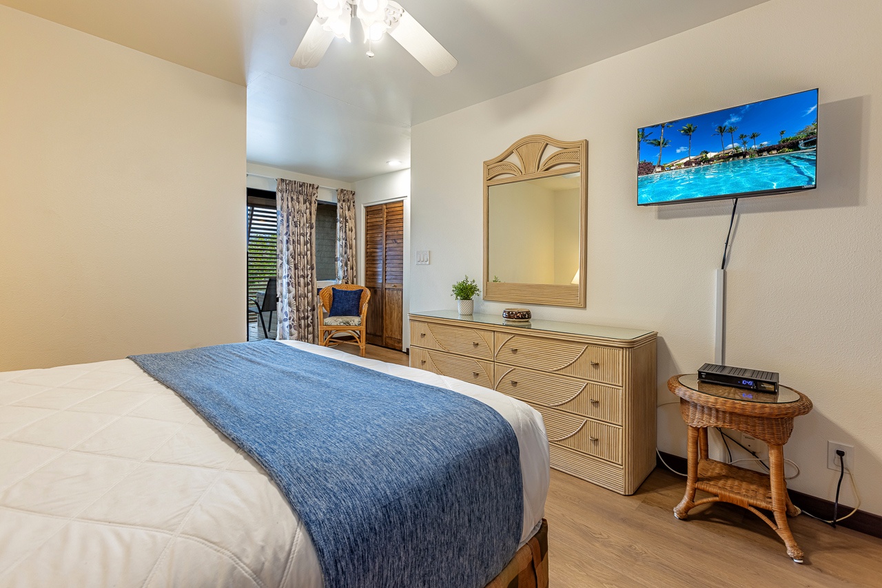 Your bright, welcoming bedroom features a cozy bed dressed in crisp linens with a cool blue accent throw, warm wood furnishings, and a ceiling fan overhead. Step through the doors onto your private outdoor space.