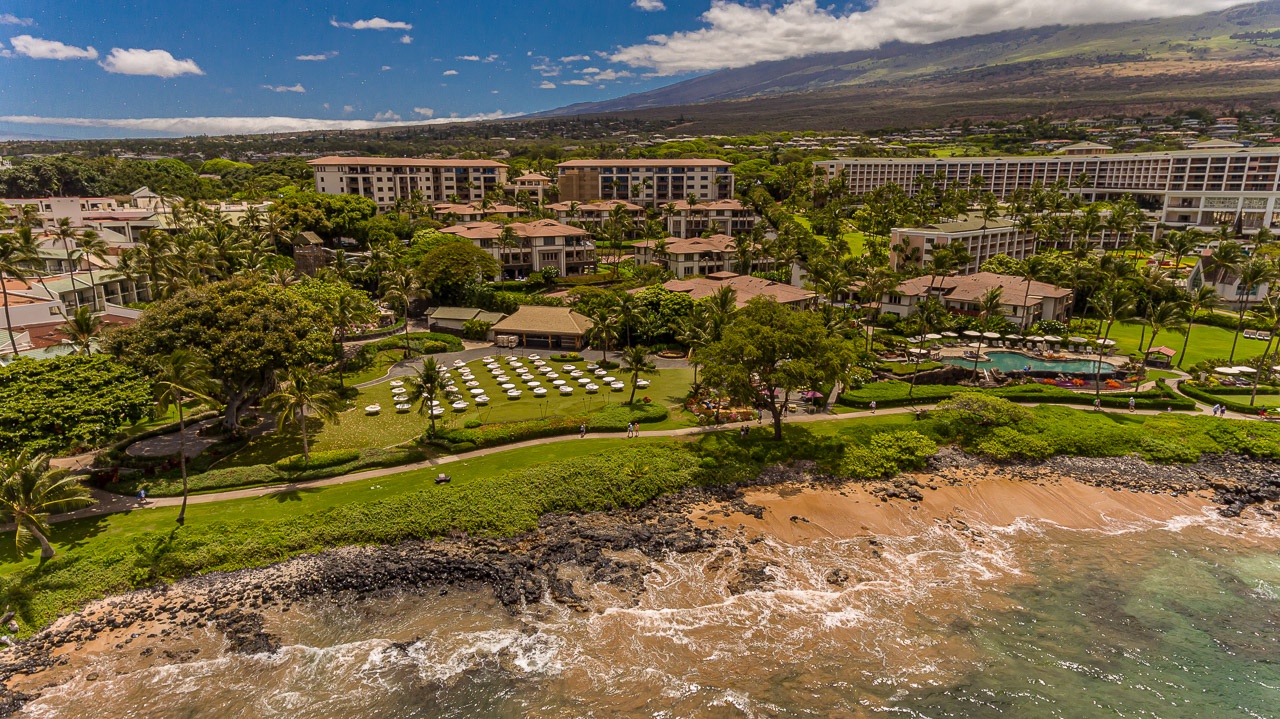 Aerial view of a beachfront resort property with lush tropical gardens, swimming pool, and sandy beach access in a scenic Hawaiian coastal setting.