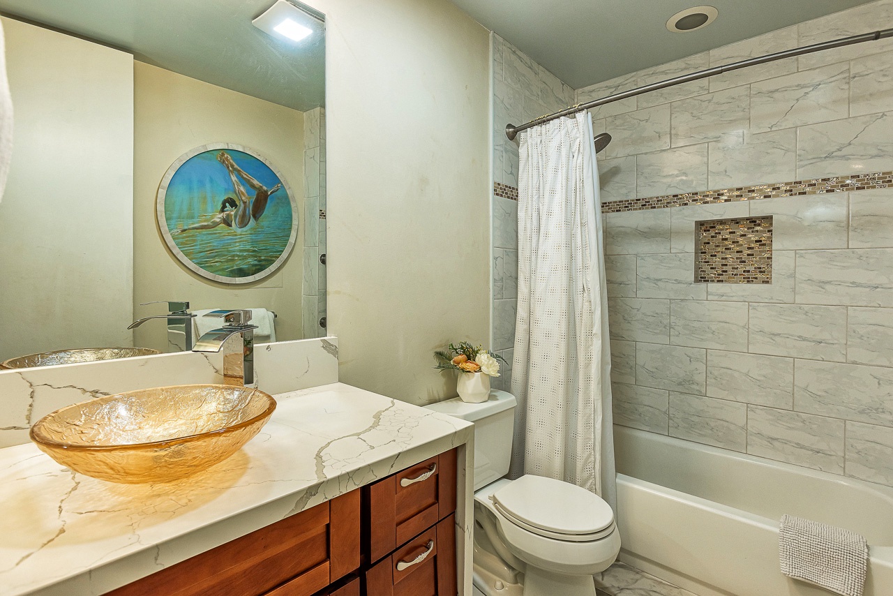 Refresh in your stylish bathroom featuring elegant marble-style tile, a luxurious glass-bowl sink, and a pristine tub-shower combo with coastal artwork welcoming you.