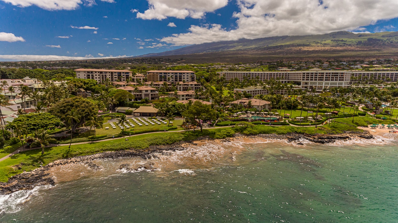 Stunning aerial perspective of the beachfront resort property nestled along Hawaii's coast, with majestic mountain landscapes rising in the distance and lush tropical gardens greeting arriving guests.