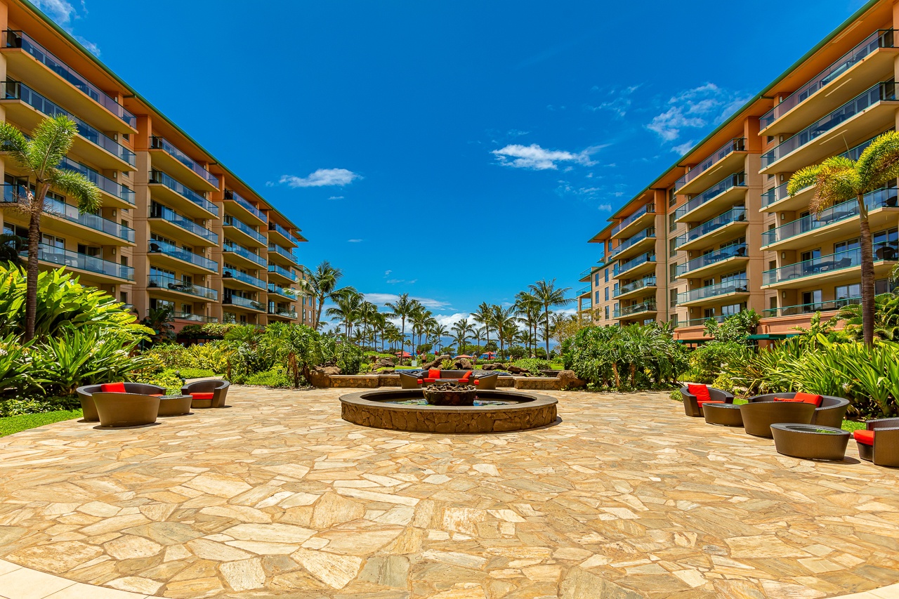 A tropical courtyard welcomes guests with a bubbling fountain, vibrant seating, and lush gardens framed by twin resort buildings under a brilliant blue sky.