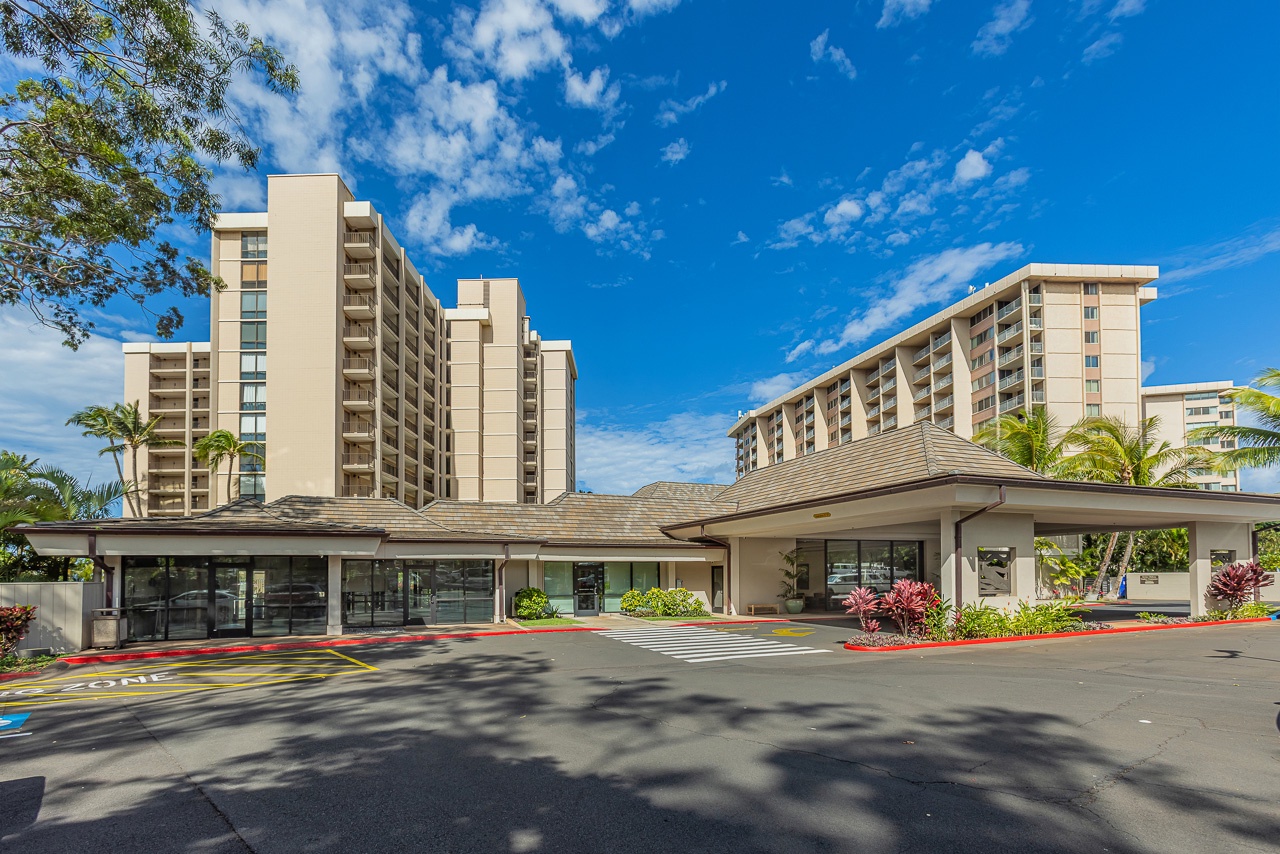 The property features a welcoming entrance with tropical landscaping and convenient covered access, set against the backdrop of its modern multi-story buildings.