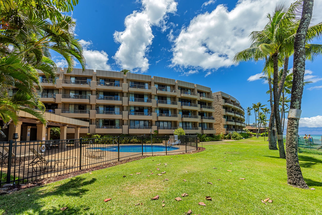 A beachfront resort building with private balconies and a refreshing pool, surrounded by swaying palm trees and lush green lawns overlooking the sparkling ocean beyond.