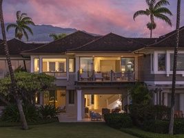 Tropical villa at dusk with mountain backdrop, palm trees, and warm interior lighting creating an inviting Hawaiian atmosphere.