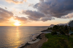 Stunning beachfront location with golden sunset views over the ocean, palm trees, and nearby resort buildings creating a tropical paradise setting.