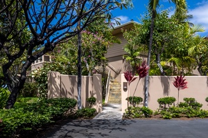 Tropical property entrance featuring lush palm trees, vibrant flowering plants, and a welcoming stone pathway leading to the main building.