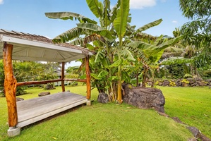 Lush tropical gardens with mature palms, decorative lava rock features, manicured lawn, and a charming thatched-roof daybed pavilion offering shade and relaxation.