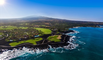 Bird's-eye view of Hawaii's coastline meeting the golf course with turquoise ocean waters beneath volcanic peaks.