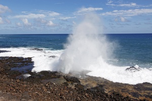 Dramatic ocean waves crash against rocky coastline, creating spectacular water geysers under bright blue skies.