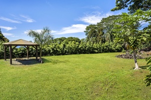Expansive green lawn bordered by lush tropical vegetation and mature trees, with a covered pavilion structure offering shaded outdoor space. The property sits beneath clear blue skies in a verdant, peaceful setting.