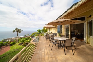 Spacious ocean-view terrace with dining table and umbrellas overlooking tropical coastline and palm trees.