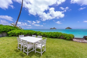 This oceanfront retreat showcases a perfectly manicured lawn with outdoor dining for six, framed by lush tropical landscaping and turquoise waters stretching toward distant islands beneath blue skies.