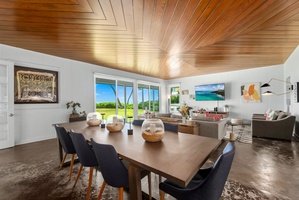 A stunning wood-paneled ceiling draws your eye upward in this open-concept living and dining space, where natural light streams through floor-to-ceiling windows revealing lush green views beyond.