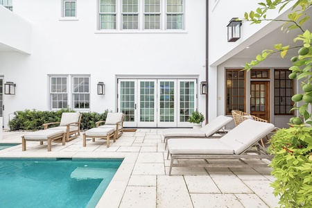 Pristine pool deck with cushioned loungers and French doors opening to interior spaces, framed by lush greenery and elegant outdoor lighting.