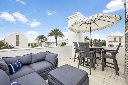 This rooftop terrace offers a private outdoor retreat within a charming resort setting, featuring a shaded dining area and comfortable lounge seating surrounded by white architecture and palm trees.