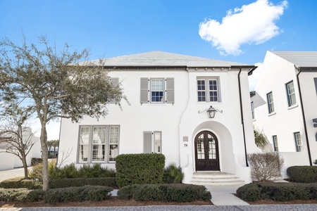 Elegant white Mediterranean-style property with charming arched entrance and manicured landscaping on a quiet cobblestone street.