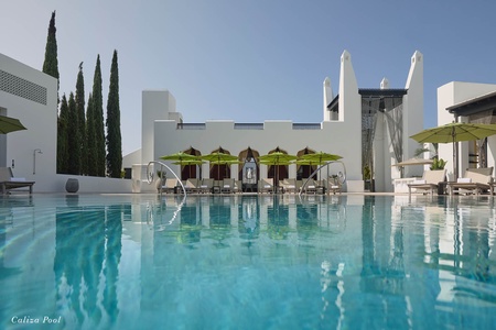 The resort features a stunning outdoor pool framed by elegant white architecture, vibrant umbrellas, and tall cypress trees—a perfect backdrop for a relaxing stay.