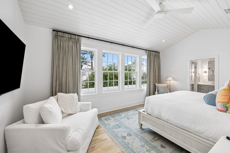 Wake in this bright, serene bedroom with vaulted shiplap ceilings, plush bedding, and a cozy reading nook, all bathed in natural light from expansive windows showcasing tropical palm views.