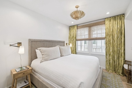 Sink into crisp white linens beneath an elegant chandelier, where golden curtains frame your city view and warm lighting creates the perfect bedtime ambiance.