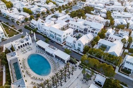 Aerial view of a beautiful residential community featuring pristine white buildings, tree-lined streets, and a stunning central pool area with restaurant facilities.