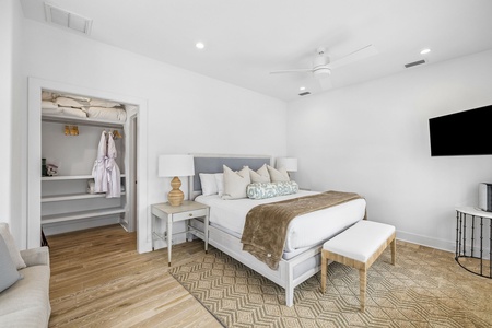 Your serene bedroom retreat features a plush bed dressed in soft linens, warm wood floors, and a mounted flat-screen TV, with a well-organized closet ready for your belongings.