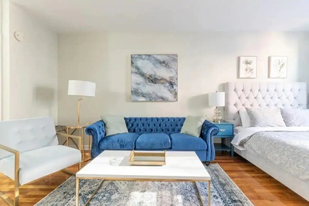 Unwind in your elegant studio where luxury meets comfort—plush blue velvet sofa, pristine white linens, and golden accents create pure sophistication.